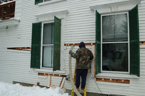 Installer on a ladder blowing cellulose insulation through holes in a clapboard wall in winter