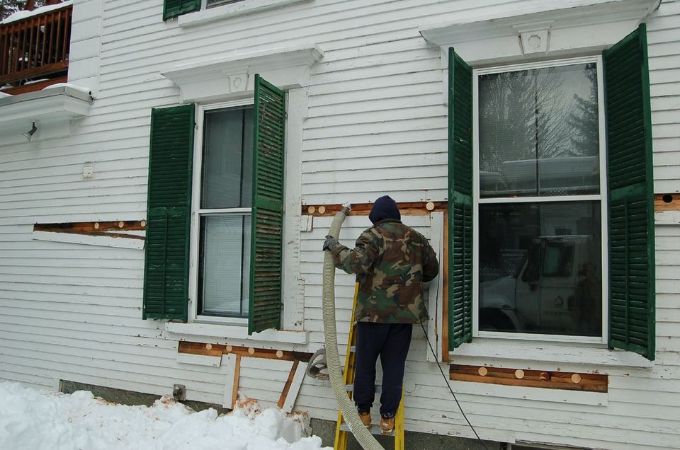 Installer on a ladder blowing cellulose insulation through holes in a clapboard wall in winter