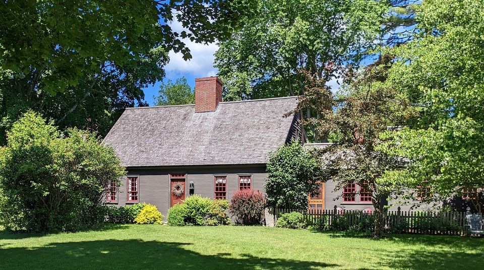 Weathered Cape Cod house with a center chimney, red trim and a picket fence