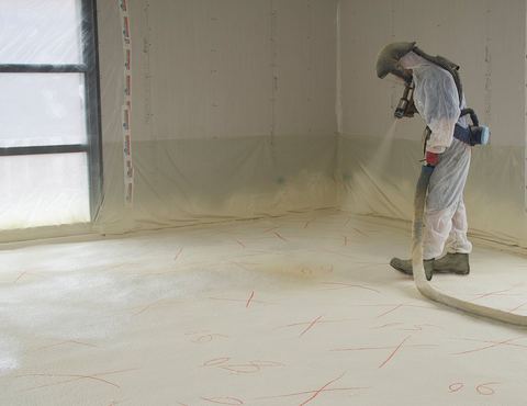 Installer in a protective suit and hood spraying foam insulation across a floor